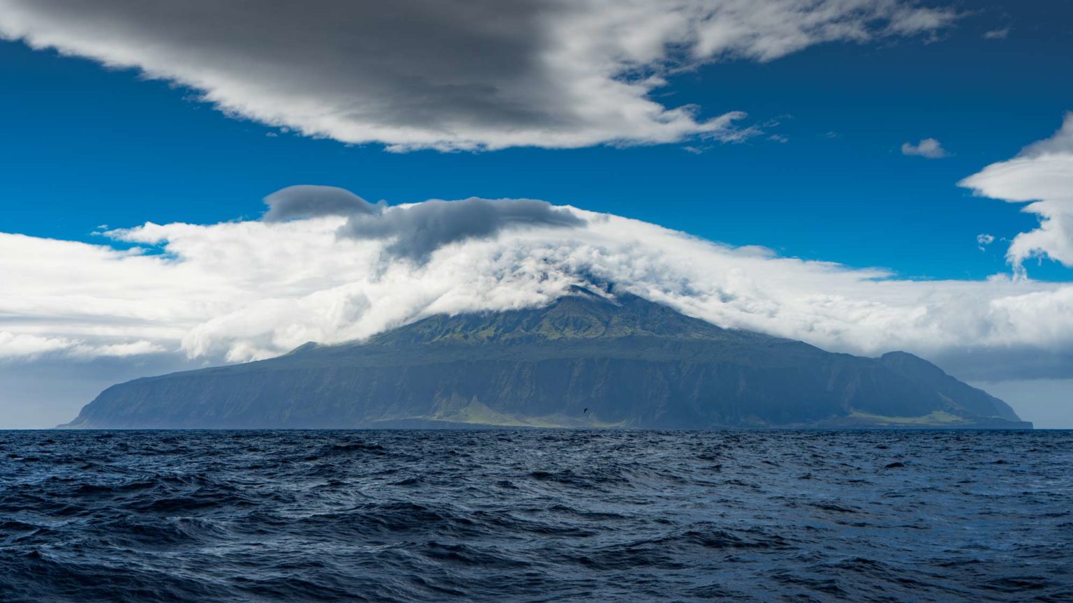 Moody analog photograph of a cloud-topped island set above a deep blue sea, atmospheric and grand.